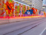 A wall lined with colorful graffiti art featuring abstract shapes and vibrant designs. The foreground shows light trails of vehicles passing by, creating a dynamic sense of movement. The backdrop includes tall urban buildings, enhancing the cityscape ambiance.