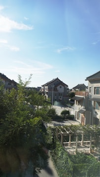 A residential area with traditional-style houses featuring gabled roofs. The area is lush with greenery, including several large trees and neatly trimmed bushes. The sky is clear, with a bright and sunny atmosphere.