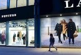 The scene depicts a storefront with mannequins dressed in stylish clothing under bright lights. Adjacent to the storefront, a large advertisement features three women walking together, dressed in fashionable coats. A single person is captured walking past on the sidewalk.