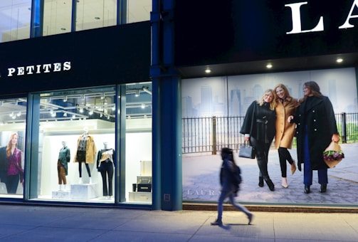 The scene depicts a storefront with mannequins dressed in stylish clothing under bright lights. Adjacent to the storefront, a large advertisement features three women walking together, dressed in fashionable coats. A single person is captured walking past on the sidewalk.