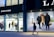 The scene depicts a storefront with mannequins dressed in stylish clothing under bright lights. Adjacent to the storefront, a large advertisement features three women walking together, dressed in fashionable coats. A single person is captured walking past on the sidewalk.