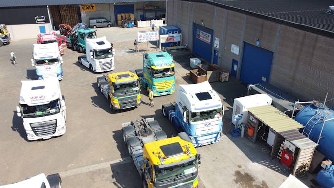 Several large trucks are parked in an industrial area next to a building with brick walls and blue doors. The trucks are mainly white with some having colorful liveries. There are signs for 'Truck Wash' and 'Exit' visible. The area is well-organized with containers and other storage facilities nearby, and a few people are present.