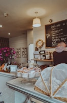 A cozy café setting with a display of pastries under a glass dome on the counter, along with flowers in a vase. The wall has a chalkboard with a menu written on it, and a person is visible at the counter.
