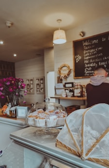 A cozy café setting with a display of pastries under a glass dome on the counter, along with flowers in a vase. The wall has a chalkboard with a menu written on it, and a person is visible at the counter.