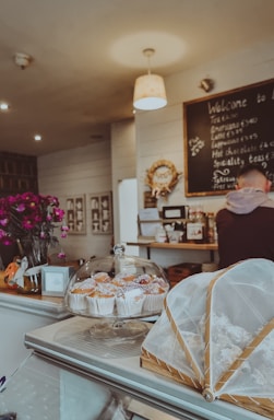 A cozy café setting with a display of pastries under a glass dome on the counter, along with flowers in a vase. The wall has a chalkboard with a menu written on it, and a person is visible at the counter.