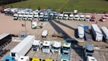An aerial view of a large parking area with numerous trucks and trailers parked in an orderly fashion. The area is divided into sections, with various white and colored trucks lined up in a few rows. A few people are walking near the trucks, and some smaller vehicles and equipment are also visible. The background includes a field with green crops.