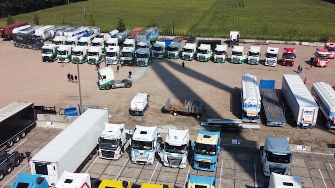 An aerial view of a large parking area with numerous trucks and trailers parked in an orderly fashion. The area is divided into sections, with various white and colored trucks lined up in a few rows. A few people are walking near the trucks, and some smaller vehicles and equipment are also visible. The background includes a field with green crops.