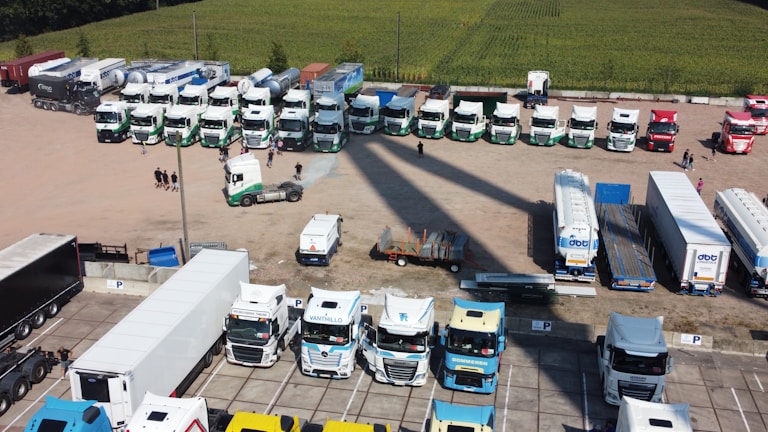 An aerial view of a large parking area with numerous trucks and trailers parked in an orderly fashion. The area is divided into sections, with various white and colored trucks lined up in a few rows. A few people are walking near the trucks, and some smaller vehicles and equipment are also visible. The background includes a field with green crops.