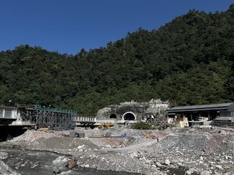 A construction site featuring a large tunnel entrance set against a forested hillside. There is a river flowing in the foreground with rocky banks. Various construction materials, scaffolding, and equipment, including vehicles and workers in safety gear, are scattered around the site. The sky is clear and blue, and the surrounding area is densely covered with trees.