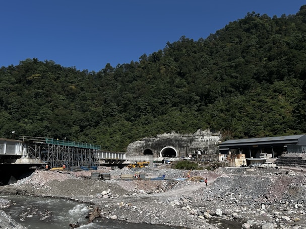 A construction site featuring a large tunnel entrance set against a forested hillside. There is a river flowing in the foreground with rocky banks. Various construction materials, scaffolding, and equipment, including vehicles and workers in safety gear, are scattered around the site. The sky is clear and blue, and the surrounding area is densely covered with trees.