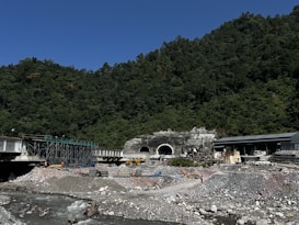 A construction site featuring a large tunnel entrance set against a forested hillside. There is a river flowing in the foreground with rocky banks. Various construction materials, scaffolding, and equipment, including vehicles and workers in safety gear, are scattered around the site. The sky is clear and blue, and the surrounding area is densely covered with trees.