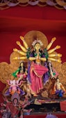 A colorful and detailed depiction of a traditional Hindu goddess with multiple arms, adorned in vibrant clothing and jewelry. The statue is part of an elaborate display that includes smaller figures, intricate patterns, and religious symbols. The background is richly decorated, enhancing the divine and festive atmosphere.