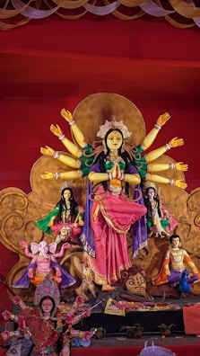 A colorful and detailed depiction of a traditional Hindu goddess with multiple arms, adorned in vibrant clothing and jewelry. The statue is part of an elaborate display that includes smaller figures, intricate patterns, and religious symbols. The background is richly decorated, enhancing the divine and festive atmosphere.