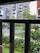 A view of a residential complex framed by bars, featuring multiple modern apartment buildings with balconies. In the foreground, lush greenery and well-maintained gardens are visible, along with a small shelter made of solar panels and a garbage bin.
