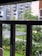 A view of a residential complex framed by bars, featuring multiple modern apartment buildings with balconies. In the foreground, lush greenery and well-maintained gardens are visible, along with a small shelter made of solar panels and a garbage bin.