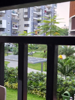 A view of a residential complex framed by bars, featuring multiple modern apartment buildings with balconies. In the foreground, lush greenery and well-maintained gardens are visible, along with a small shelter made of solar panels and a garbage bin.