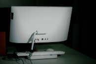 A white desktop computer monitor is seen from the back, sitting on a table with a keyboard and mouse placed in front. The setup includes some visible cables, and the background is dimly lit, creating a shadowy effect.