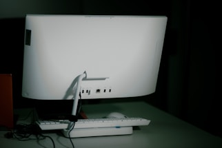 A white desktop computer monitor is seen from the back, sitting on a table with a keyboard and mouse placed in front. The setup includes some visible cables, and the background is dimly lit, creating a shadowy effect.