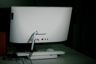 A white desktop computer monitor is seen from the back, sitting on a table with a keyboard and mouse placed in front. The setup includes some visible cables, and the background is dimly lit, creating a shadowy effect.