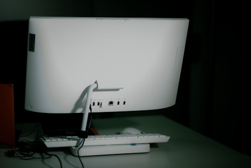 A white desktop computer monitor is seen from the back, sitting on a table with a keyboard and mouse placed in front. The setup includes some visible cables, and the background is dimly lit, creating a shadowy effect.