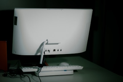 A white desktop computer monitor is seen from the back, sitting on a table with a keyboard and mouse placed in front. The setup includes some visible cables, and the background is dimly lit, creating a shadowy effect.