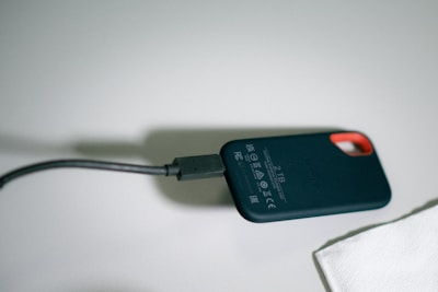 A small external solid-state drive (SSD) with a capacity label of 2 TB is connected to a black cable. The device has rounded edges and a keyhole design for a clip or lanyard. It sits on a light-colored surface with a piece of white paper or cloth nearby.