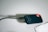 A small external solid-state drive (SSD) with a capacity label of 2 TB is connected to a black cable. The device has rounded edges and a keyhole design for a clip or lanyard. It sits on a light-colored surface with a piece of white paper or cloth nearby.