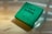 A green box labeled 'Love Bathing' with the word 'Soap' and the Hindi translation 'साबुन'. Water droplets are scattered across the box, giving it a fresh appearance. The light reflects off the surface, casting soft shadows.