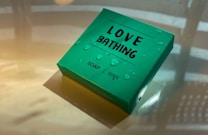 A green box labeled 'Love Bathing' with the word 'Soap' and the Hindi translation 'साबुन'. Water droplets are scattered across the box, giving it a fresh appearance. The light reflects off the surface, casting soft shadows.