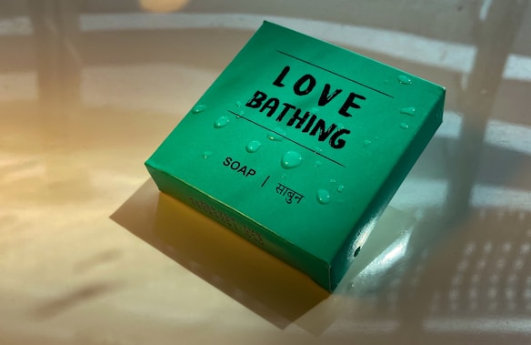 A green box labeled 'Love Bathing' with the word 'Soap' and the Hindi translation 'साबुन'. Water droplets are scattered across the box, giving it a fresh appearance. The light reflects off the surface, casting soft shadows.