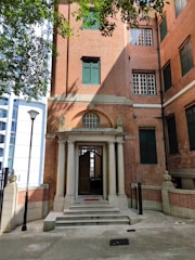 A red-brick building with a classical style entrance featuring four columns and a small set of stairs. The entryway includes a wooden door partially open, and the windows are adorned with dark green shutters. The building is flanked by lampposts, and overhead, tree branches cast shadows on the scene.