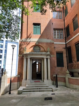 A red-brick building with a classical style entrance featuring four columns and a small set of stairs. The entryway includes a wooden door partially open, and the windows are adorned with dark green shutters. The building is flanked by lampposts, and overhead, tree branches cast shadows on the scene.