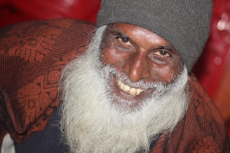 An elderly person with a long white beard and a warm smile, wearing a dark knitted beanie and a patterned shawl. The background consists of warm, blurred tones that enhance the focus on the person's joyful expression.