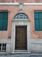 A wooden double door with decorative panels set in a brick facade. The entrance is framed by a stone arch with a semi-circular window above, divided into multiple sections by muntins. Green shutters flank the door and the bricks have a warm, reddish hue. Above the door, there is a decorative stone crest featuring stylized motifs.