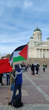 A person stands on a cobblestone plaza holding a flag with a combination of red, black, white, and green colors, commonly associated with Palestine. The individual faces a large white neoclassical building with towers and domes, surrounded by other people who appear to be gathering or walking. The sky is partly cloudy.