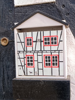 A wooden box is painted with a simple rustic house design featuring windows with red frames and black lattice patterns against a white background. Next to the box is a small round brass-colored fixture on a black and white wall.