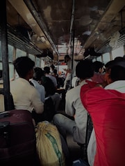 A crowded bus interior with passengers seated closely together. Luggage and personal belongings like bags are seen on the floor and overhead shelves. The bus is dimly lit, with several people engaged in conversation. The atmosphere seems busy and close-knit.
