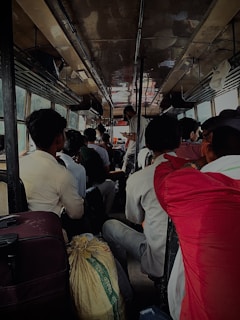 A crowded bus interior with passengers seated closely together. Luggage and personal belongings like bags are seen on the floor and overhead shelves. The bus is dimly lit, with several people engaged in conversation. The atmosphere seems busy and close-knit.