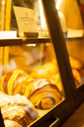 A display case filled with freshly baked pastries, including swirled and powdered varieties. The lighting is warm, casting a golden glow on the baked goods. A sign labeled 'Banana Cake' is visible inside the case.
