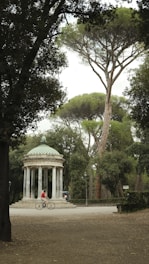 A small, classical rotunda with a domed roof and several columns stands amidst a lush, green park. A person wearing a red shirt rides a bicycle in front of the structure, and another person is seated nearby under the shade of tall trees. The scene is framed by dense foliage and natural greenery.
