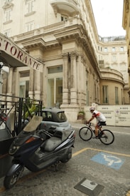 A street scene featuring a pedestrian area with a grand, historic building in the background. A person in cycling gear rides a bicycle past a scooter parked near a small, compact car. Signs indicate restrictions related to private parking, and the pavement is comprised of cobblestones.