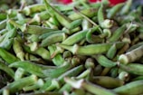 A close-up view of a pile of fresh green okra pods, displaying their unique ribbed texture and pointed tips. The okra is densely packed, creating an impression of abundance.