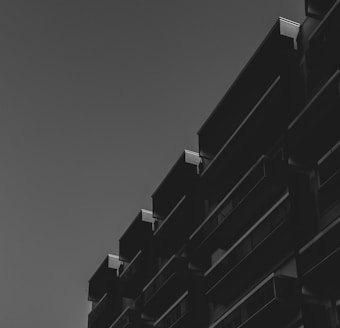 A dark, monochromatic image featuring the silhouette of an angular, modern building with protruding balconies against a clear sky. The lighting highlights the edges, creating a stark contrast between the building and the sky.
