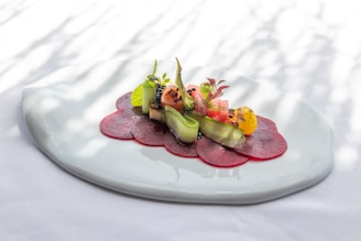 A beautifully arranged dish featuring thin slices of red beetroot topped with a variety of colorful vegetables, including cucumber ribbons, cherry tomatoes, and leafy greens. The presentation is elegant, with a focus on freshness and artistry, served on a white textured plate with a soft, natural light casting shadows.