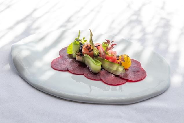 A beautifully arranged dish featuring thin slices of red beetroot topped with a variety of colorful vegetables, including cucumber ribbons, cherry tomatoes, and leafy greens. The presentation is elegant, with a focus on freshness and artistry, served on a white textured plate with a soft, natural light casting shadows.