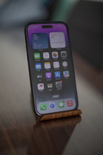 A smartphone is displayed on a wooden stand with its screen turned on. The smartphone shows various app icons and a weather widget displaying temperature and weather conditions in Berlin. The screen has a purple gradient wallpaper and is set against a blurred background, possibly a dark wooden surface.