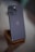 A sleek smartphone with a metallic finish stands on a wooden stand. The phone has three prominent camera lenses on the back, along with a small flash. The setting is minimalistic, with a soft blurred background indicating a modern, sophisticated ambiance.