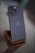 A sleek smartphone with a metallic finish stands on a wooden stand. The phone has three prominent camera lenses on the back, along with a small flash. The setting is minimalistic, with a soft blurred background indicating a modern, sophisticated ambiance.