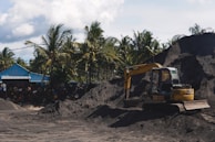 A yellow excavator is positioned on a large pile of dark soil in a yard surrounded by tall palm trees. Next to it, a blue structure with a sloped roof is visible, alongside a stack of weathered barrels. The sky is partly cloudy, and the scene conveys an industrial setting amidst a tropical environment.