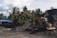 A yellow excavator is positioned on a large pile of dark soil in a yard surrounded by tall palm trees. Next to it, a blue structure with a sloped roof is visible, alongside a stack of weathered barrels. The sky is partly cloudy, and the scene conveys an industrial setting amidst a tropical environment.